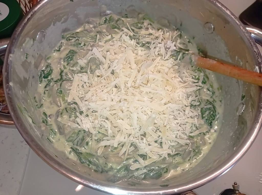 Grated Parmesan being added to a pan of sautéed spinach and mushrooms in creamy sauce