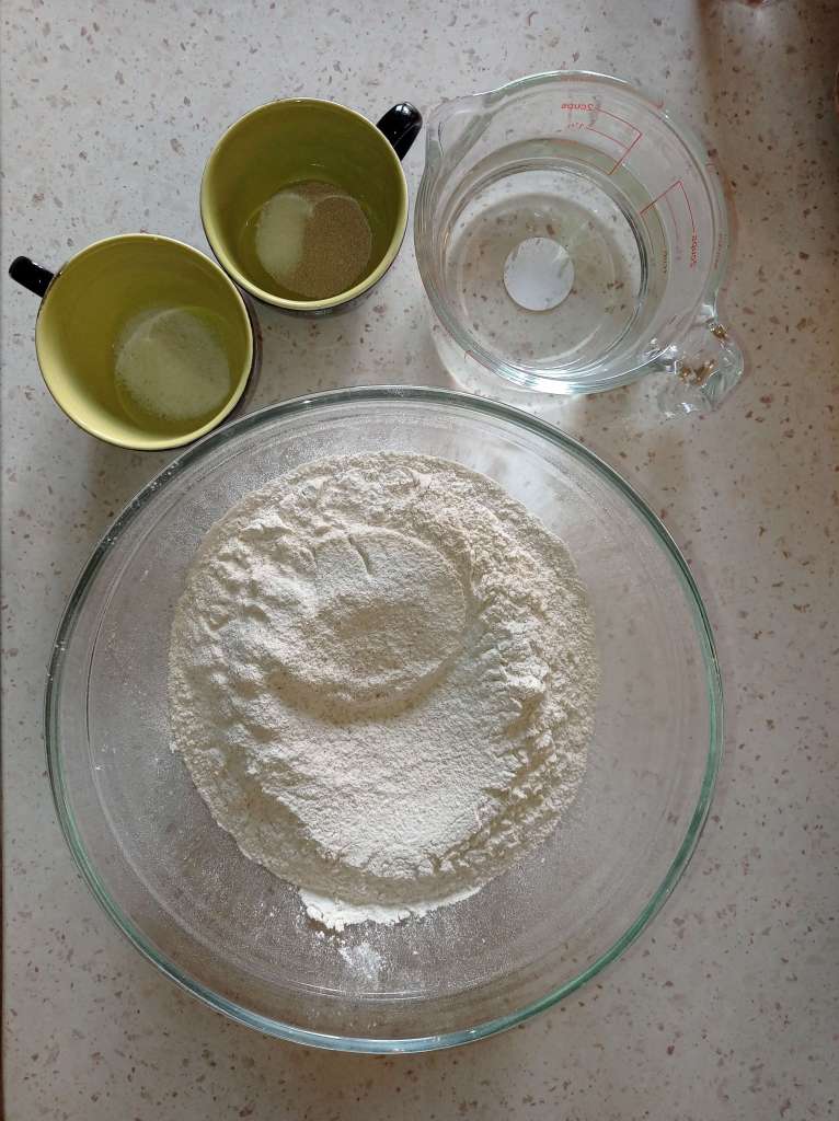 Ingredients for homemade bread: cup of salt, cup of yeast and sugar, bowl of water, and large bowl of flour.