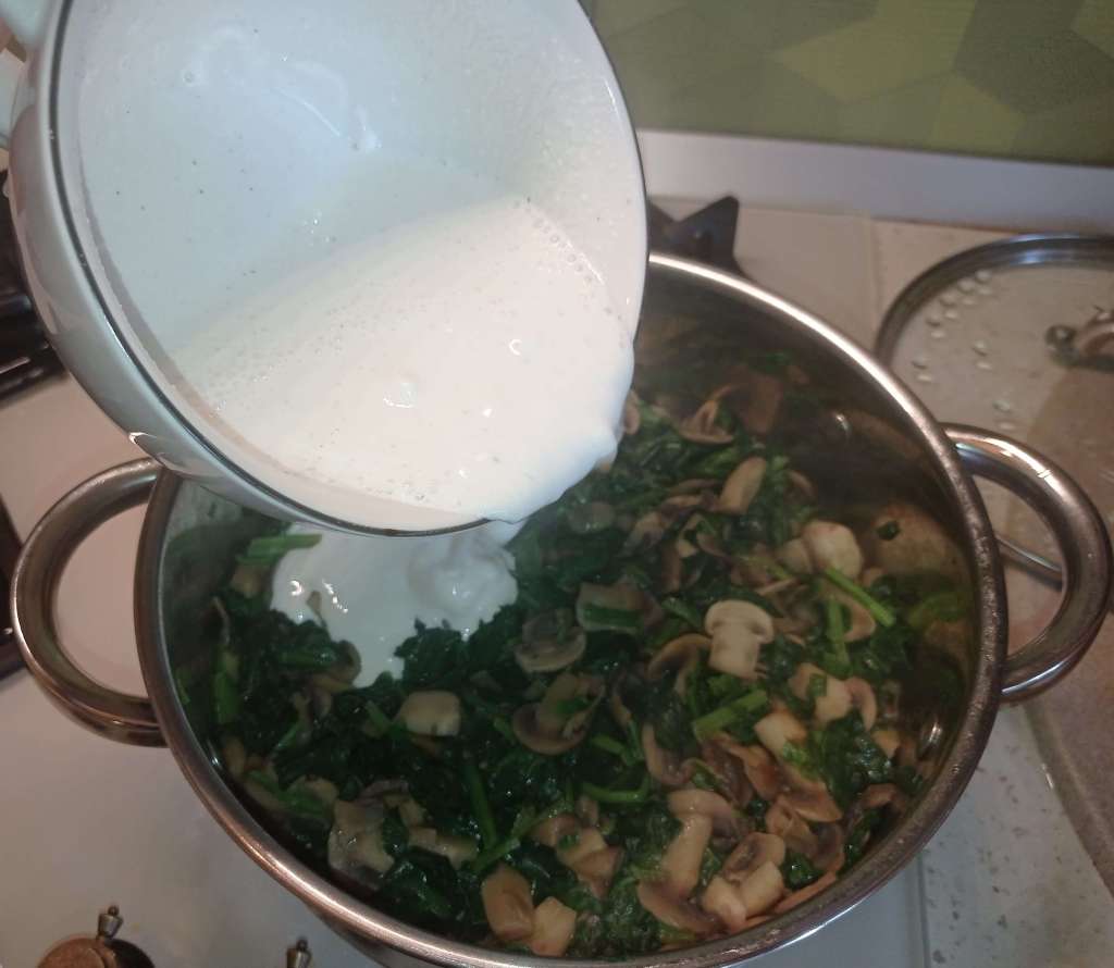 Pouring milk, sour cream, and starch mixture into a skillet with sautéed spinach and mushrooms