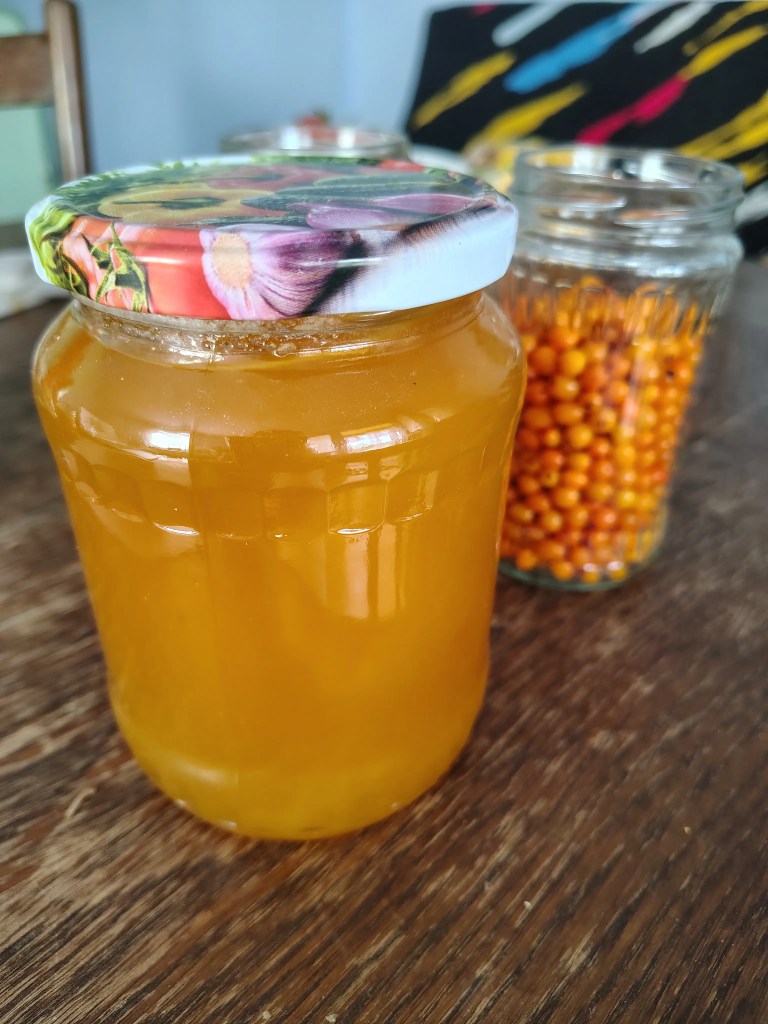 A jar filled with golden honey next to glass jars of fresh sea buckthorn berries on a rustic wooden table, ready to be combined for the maceration process.